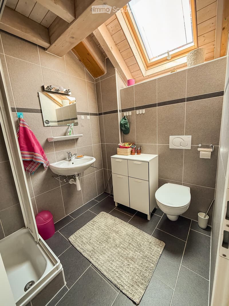 Modern bathroom with grey tiles, white sink, toilet, cabinet, mirror, and a small window with wooden frame.