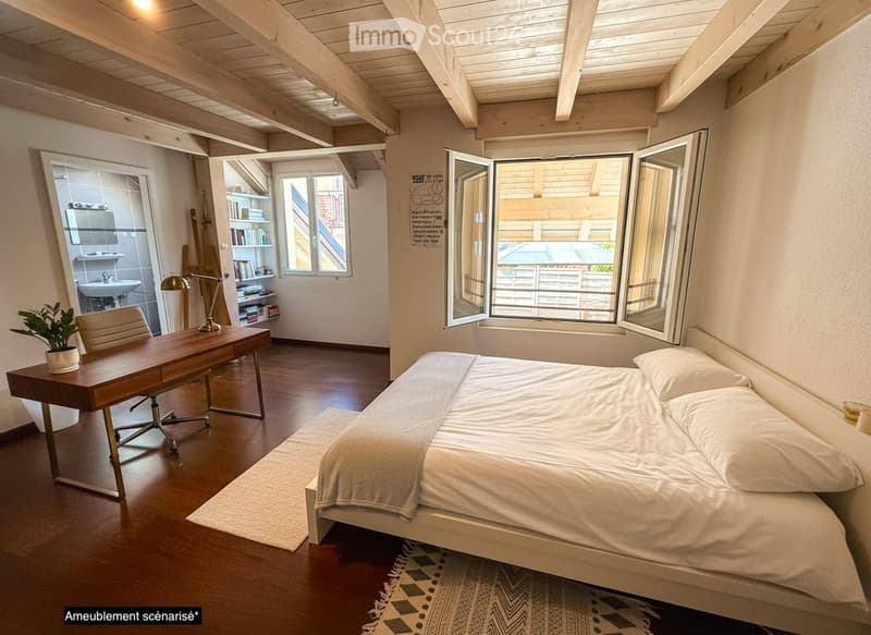 Bright, modern bedroom with a large bed, wooden desk, chair, lamp, and a small plant. The room features exposed wooden beams on the ceiling and large windows with open shutters, allowing natural light to fill the space. A small bathroom is visible through