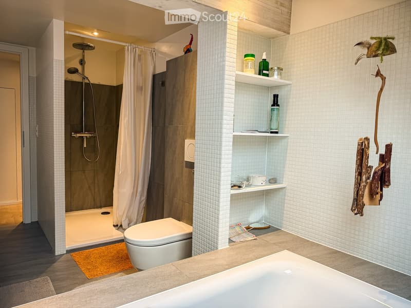 Modern bathroom with a white toilet, bathtub, and shower area. Shelves hold various toiletries and decorative items, with a small rug placed near the toilet.