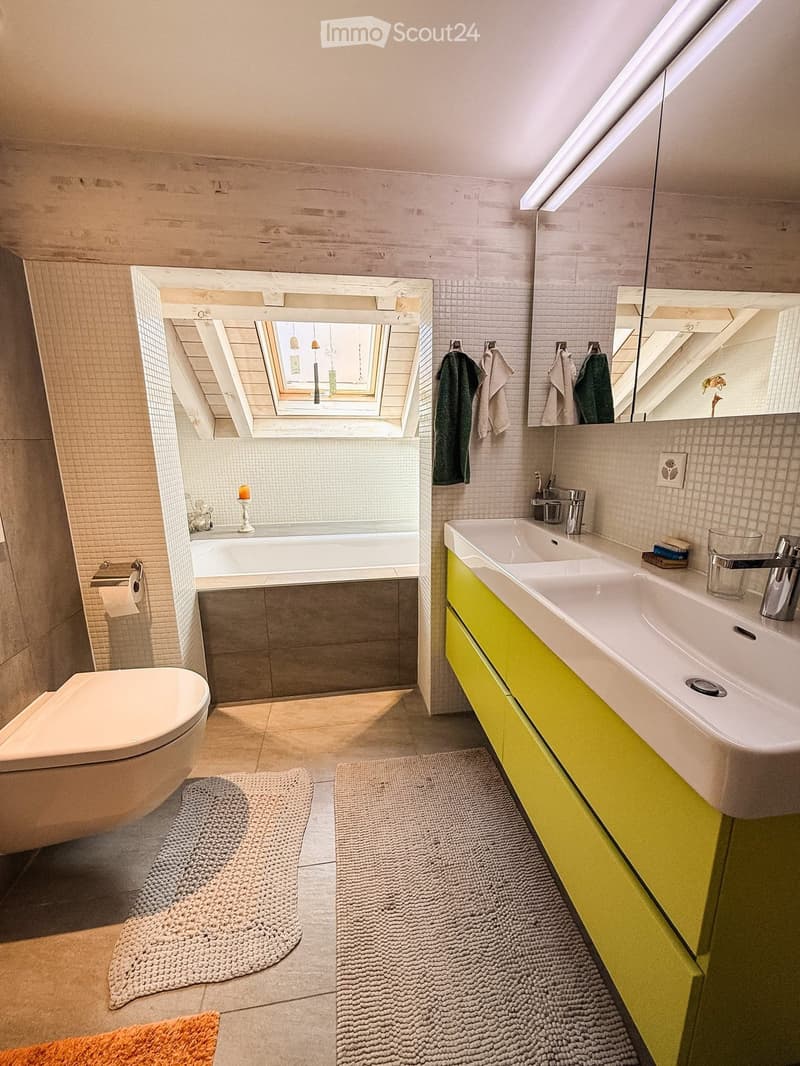 Modern bathroom with a bathtub, toilet, double sink, and large mirror. Features include a skylight window, bright yellow cabinets, and two round bath mats.
