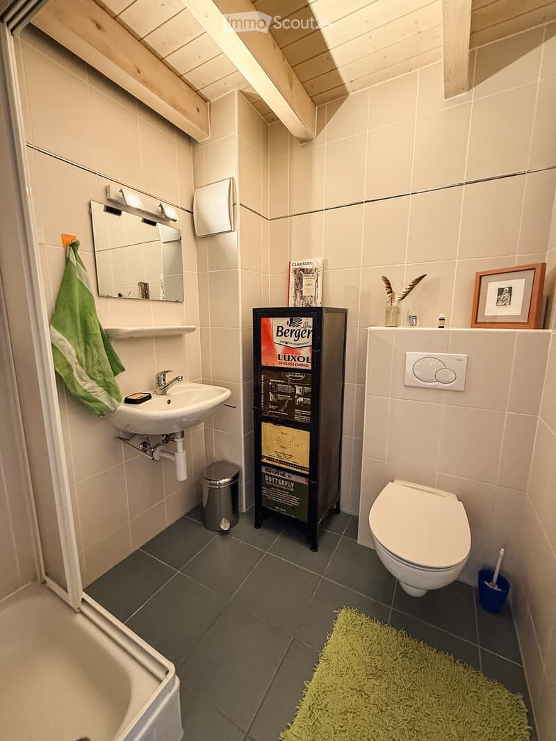 Modern bathroom with white tiles, a sink, mirror, toilet, and a small green bath mat. A towel hangs on the wall, and a small cabinet holds various items.