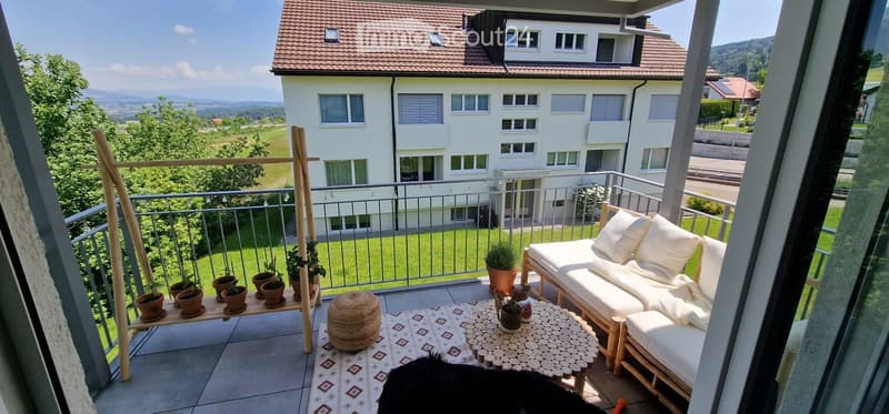 Balcony with a view of a residential building and green landscape, featuring a white sofa, small round table, potted plants, and a woven rug.