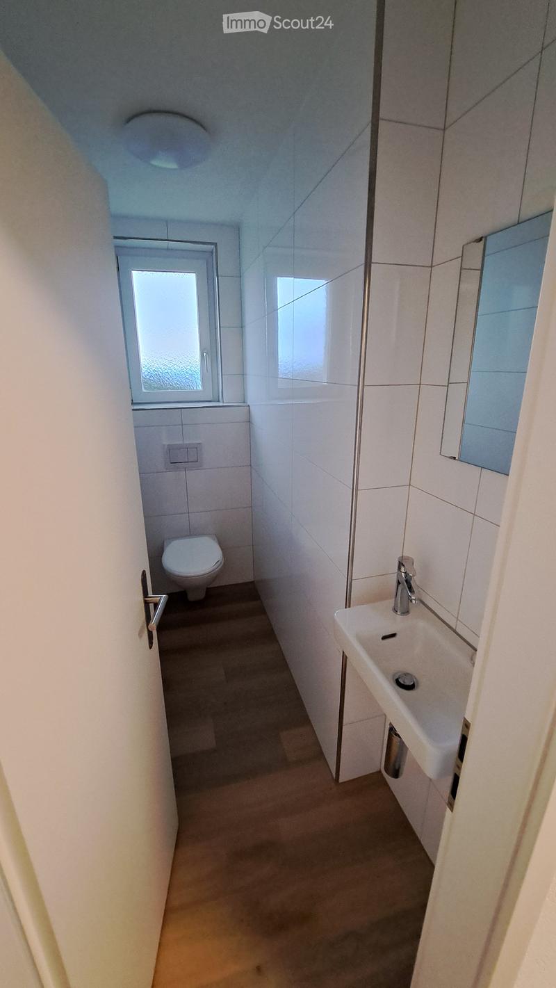 Modern bathroom with white tiles, a toilet, a sink with a faucet, and a mirror. The space is compact with a small window providing natural light.