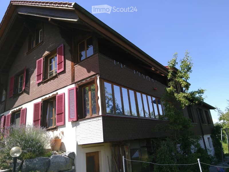 Two-story house with red shutters, large windows, and a balcony on the upper level. The house features a mix of brick and wood exterior, surrounded by greenery and a stone retaining wall.