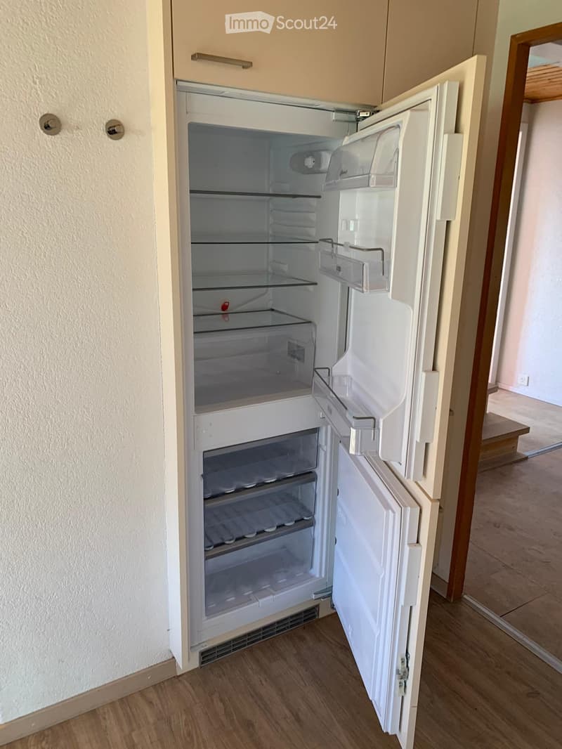 Empty white refrigerator with multiple shelves and compartments, located in a room with wooden flooring and light-colored walls.