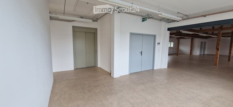Empty room with white walls, concrete floor, and two doors. Exposed wooden beams and industrial-style lighting fixtures are visible in the background.