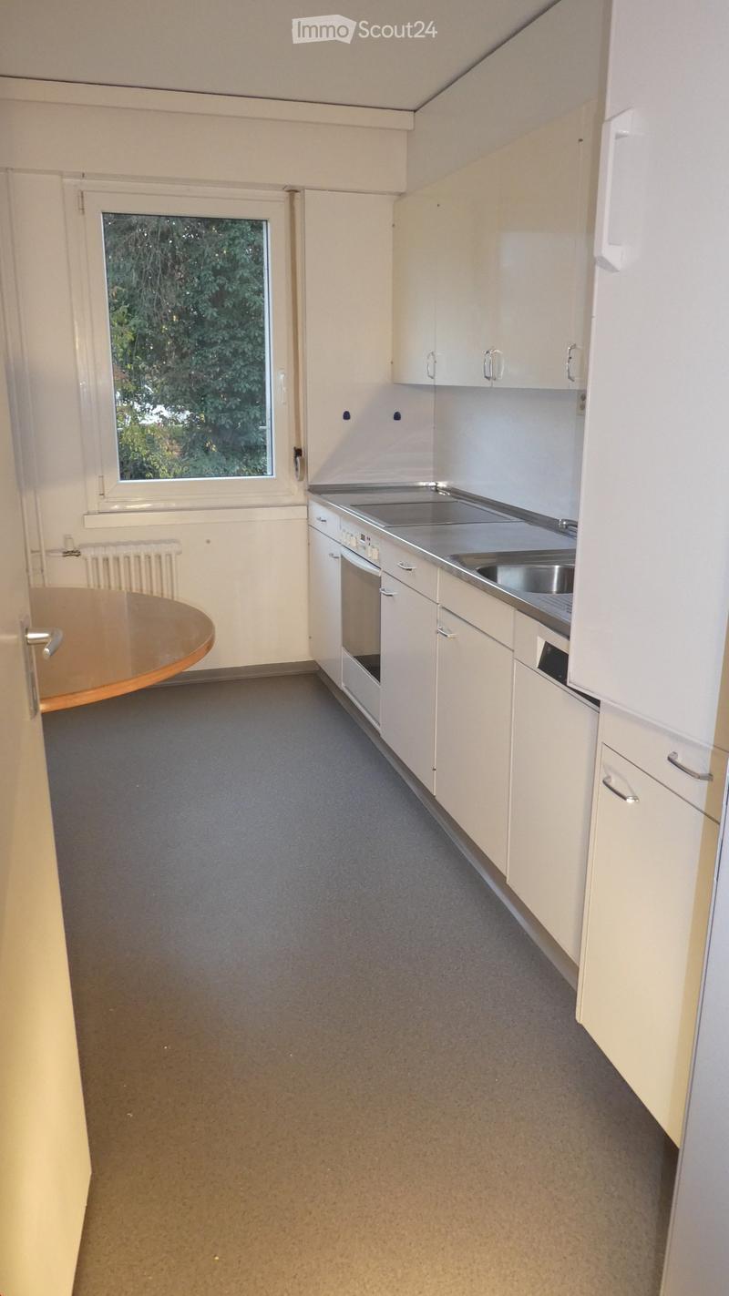 The kitchen has a white refrigerator, a gas stove, a wooden table, and a window with a white frame. The wall is white and there are white cabinets. There are two power outlets on the wall. On the wall, there is a radiator and a faucet. The floor is tiled.