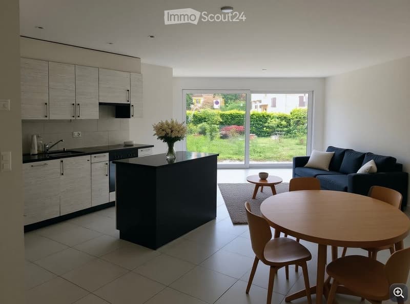 Modern open-plan living and dining area with a kitchen, featuring a dark island, light grey cabinets, a blue sofa, a round wooden dining table with chairs, and a small round side table with a bowl.