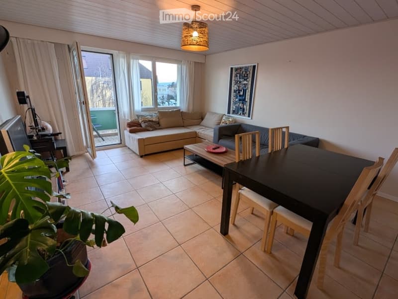 Open-plan living and dining area with a sectional sofa, coffee table, dining table with chairs, and a potted plant near the entrance to a balcony.