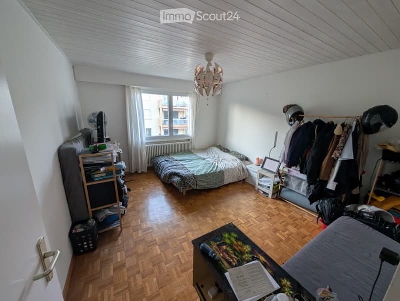 A small, furnished bedroom with wooden flooring, a bed, a table, a chair, a fan, a screen, a radiator, a curtain, a lamp, and a wardrobe with shelves.