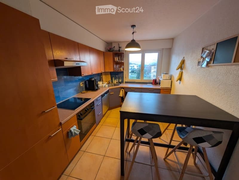 Modern kitchen with wooden cabinets, stainless steel appliances, a central island table with two stools, and a window offering a view.