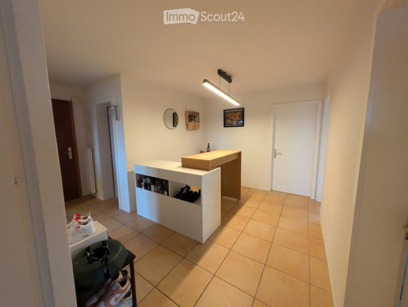 A modern, minimalist hallway with a built-in bar counter, a round mirror, framed picture, and shelves holding shoes and luggage.