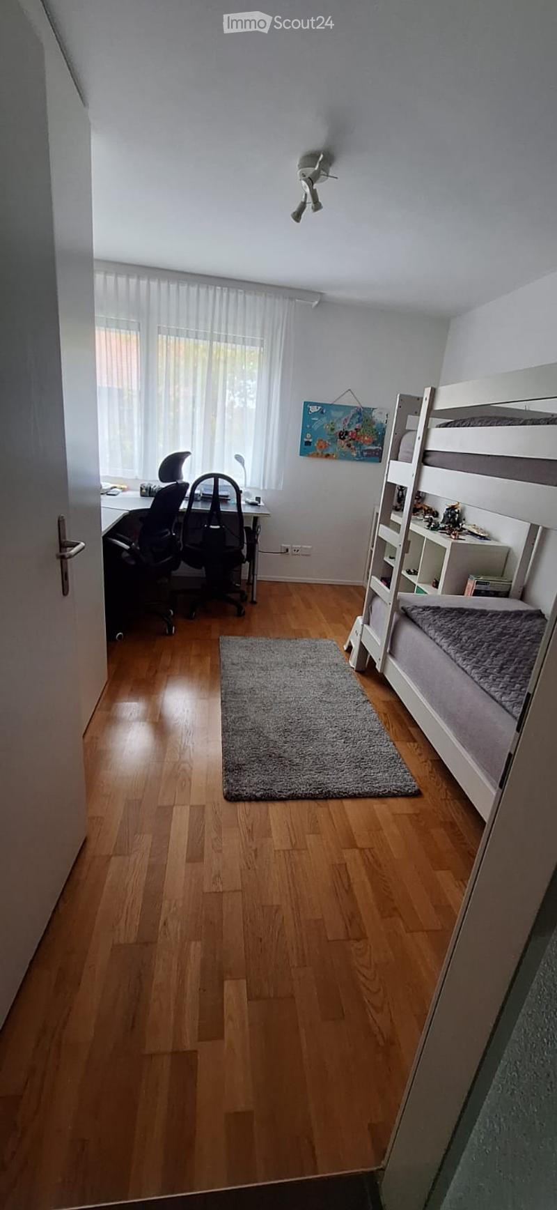 A well-lit bedroom with wooden flooring, featuring a bunk bed, a desk with a chair and laptop, and a gray rug on the floor.