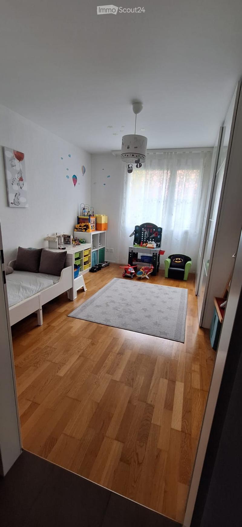 A well-lit children's bedroom with wooden flooring, a white sofa, a Batman chair, a small table with toys, a shelf with colorful storage boxes, and a hanging lamp.