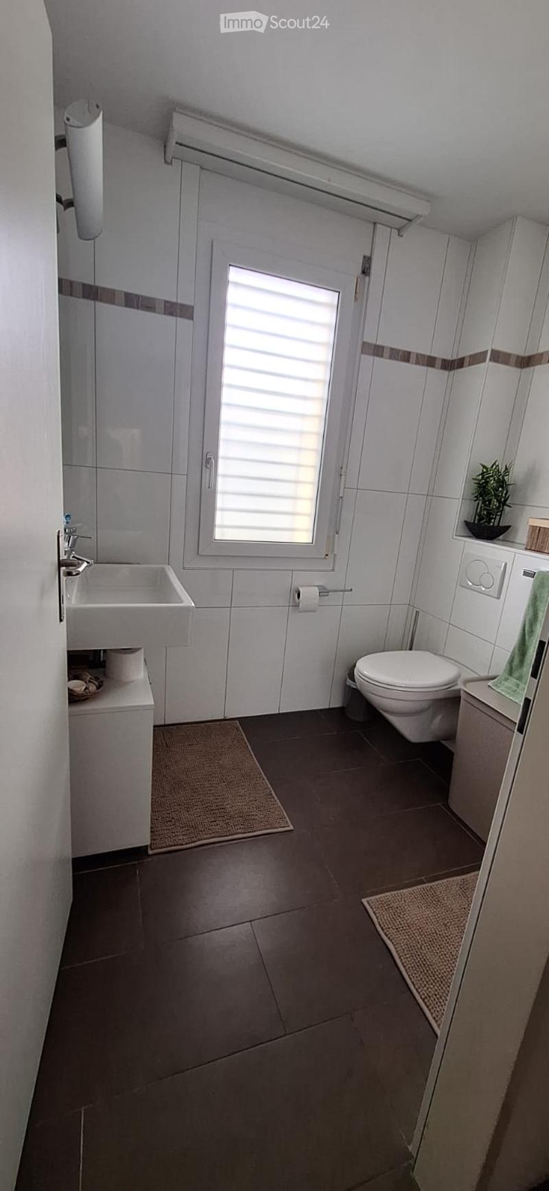 Modern bathroom with white tiles, dark brown floor tiles, a window with blinds, a sink with a faucet, a toilet, and a small plant on the shelf.