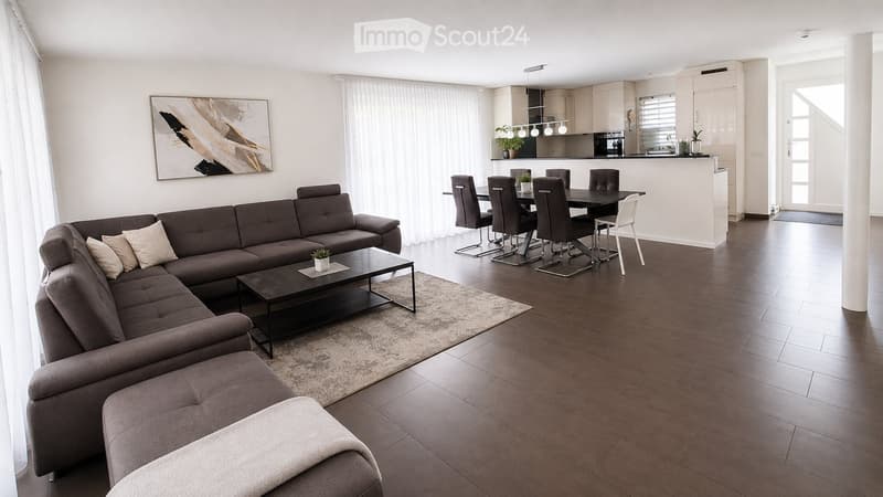 Modern living and dining area with a sectional sofa, coffee table, ottoman, dining table with chairs, and a kitchen area with cabinets and pendant lighting.