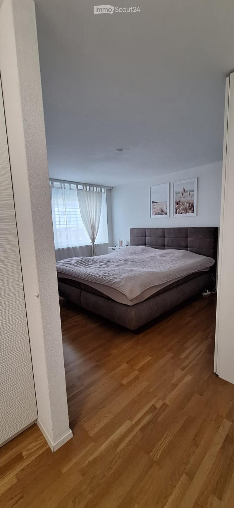 Modern bedroom with wooden flooring, large bed with grey headboard, nightstand, and two picture frames on the wall.