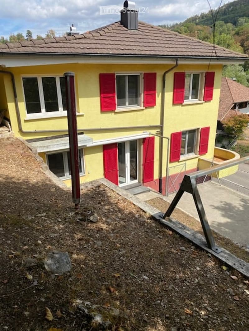 Two-story yellow house with red shutters, white-framed windows, and a tiled roof. The house has a raised ground floor and is surrounded by a gravel area.