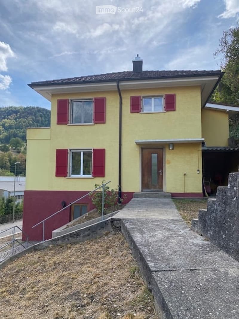 Two-story house with yellow and red exterior, red shutters, and a raised ground floor accessed by concrete steps.