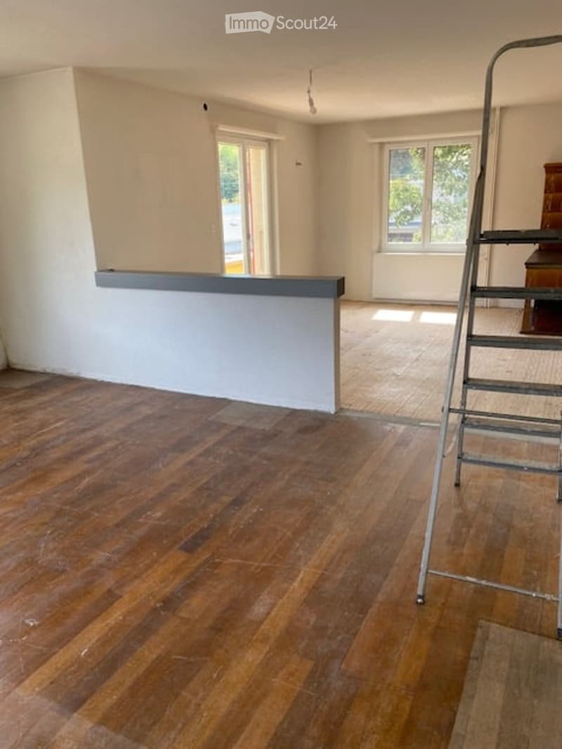Empty room with wooden flooring, white walls, and large windows allowing natural light. A small built-in bench or counter is visible along one wall.
