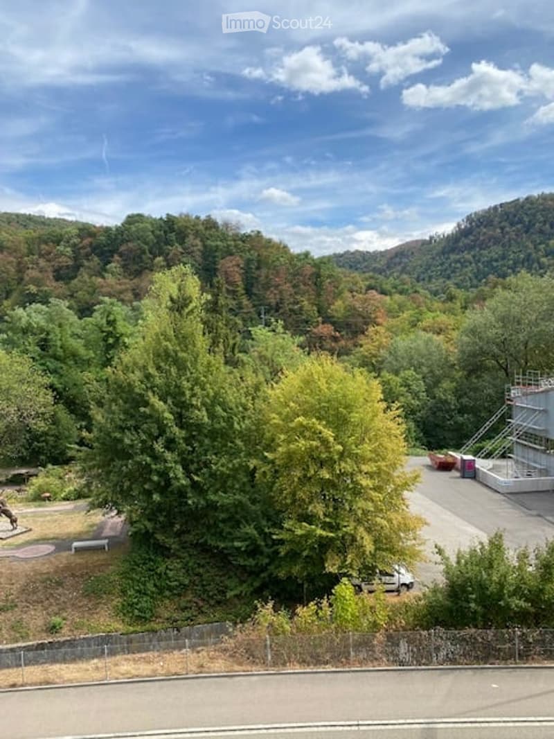 A park-like outdoor area with trees, a paved pathway, and a small structure, likely a playground or shelter, surrounded by greenery and hills in the background.