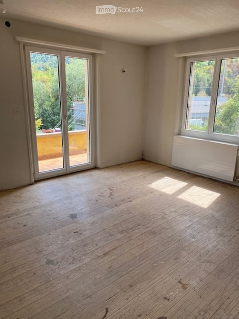 Empty room with wooden flooring, large sliding glass door leading to a balcony, and a window with a radiator below.
