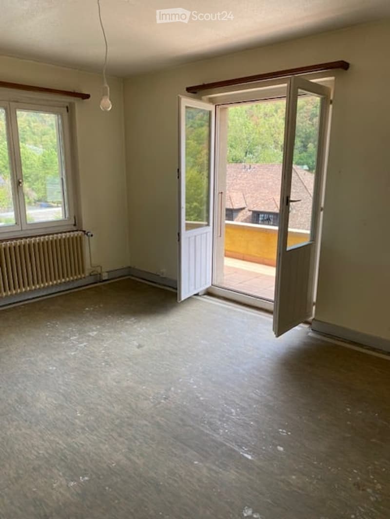 Empty room with wooden flooring, large window, open door leading to a balcony or terrace, radiator on the wall, and visible light fixture.