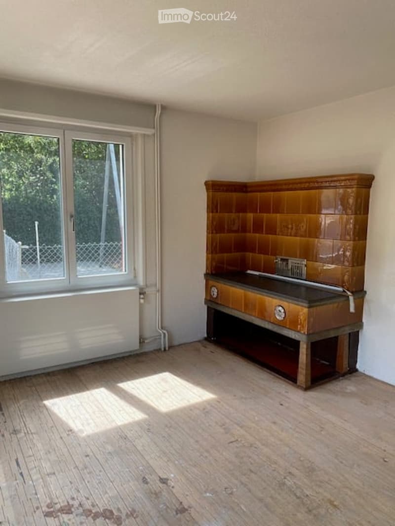 Empty room with wooden flooring, large window with white frame, and a built-in tiled stove with two dials.