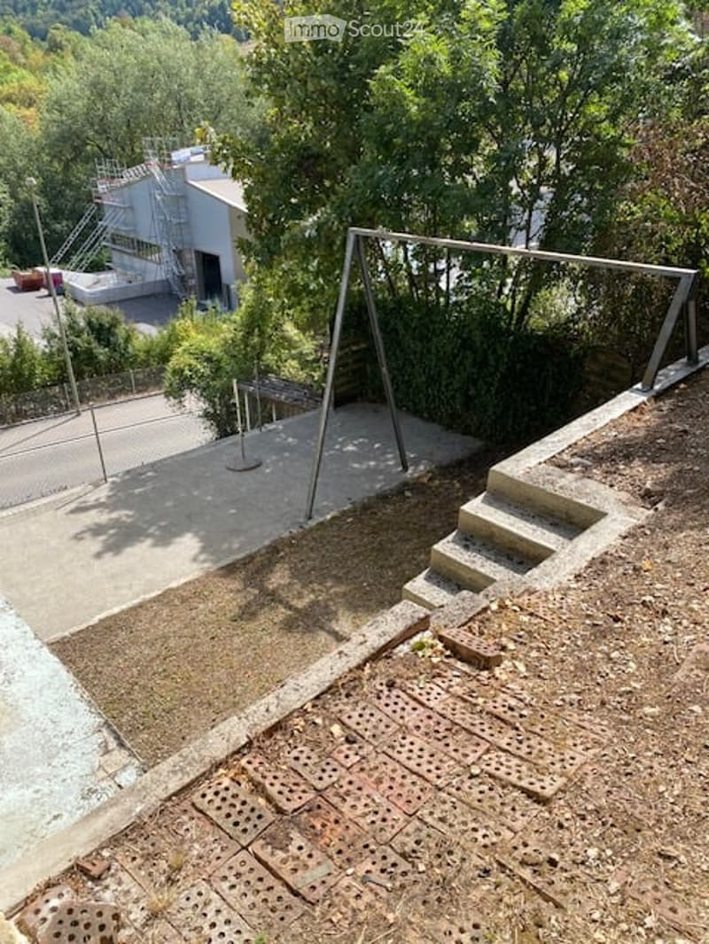An outdoor area with a raised ground floor, concrete steps leading up to a platform, and a metal structure, likely part of a playground or recreational area.