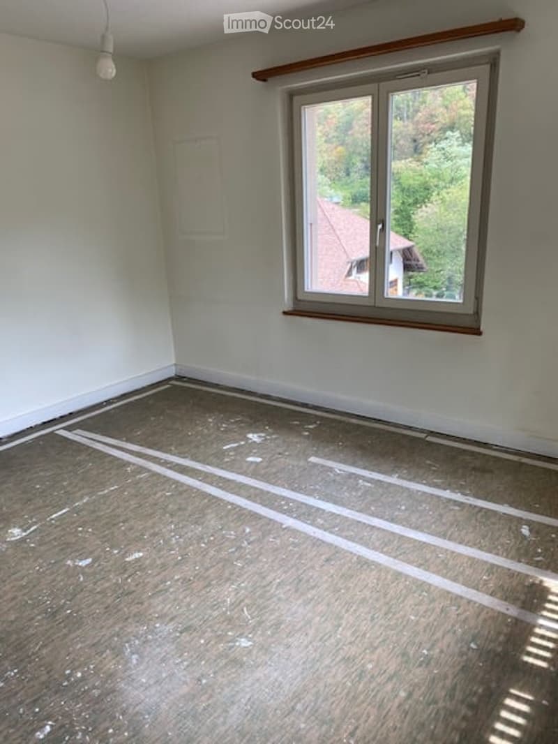 Empty room with bare walls, wooden floor, and a large window showing a view of trees and a neighboring house.