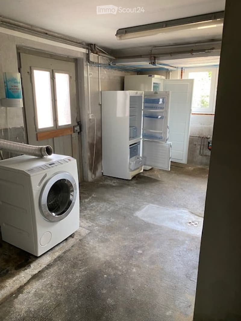 Empty room with a washing machine and an open refrigerator, concrete floor, exposed pipes, and windows.