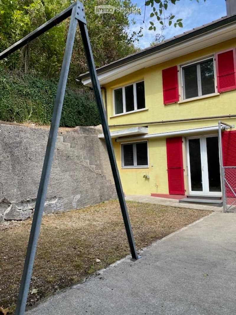 Two-story yellow house with red shutters, raised ground floor, and a concrete retaining wall.