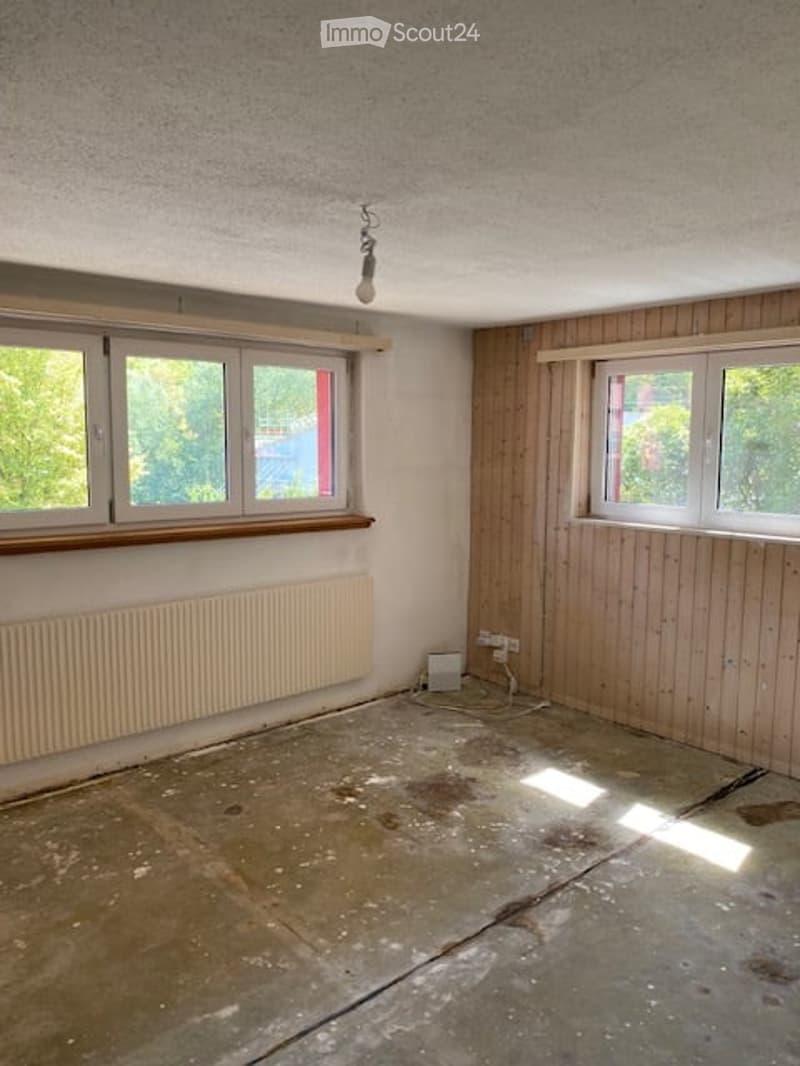 Empty room with wooden panel walls, concrete floor, and large windows with wooden frames. Basic electrical outlets and a radiator are visible.