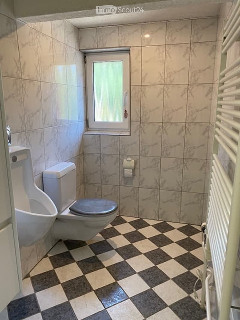 Modern bathroom with marble tiles, a toilet, urinal, and a small window. The floor features a classic black and white checkered pattern.