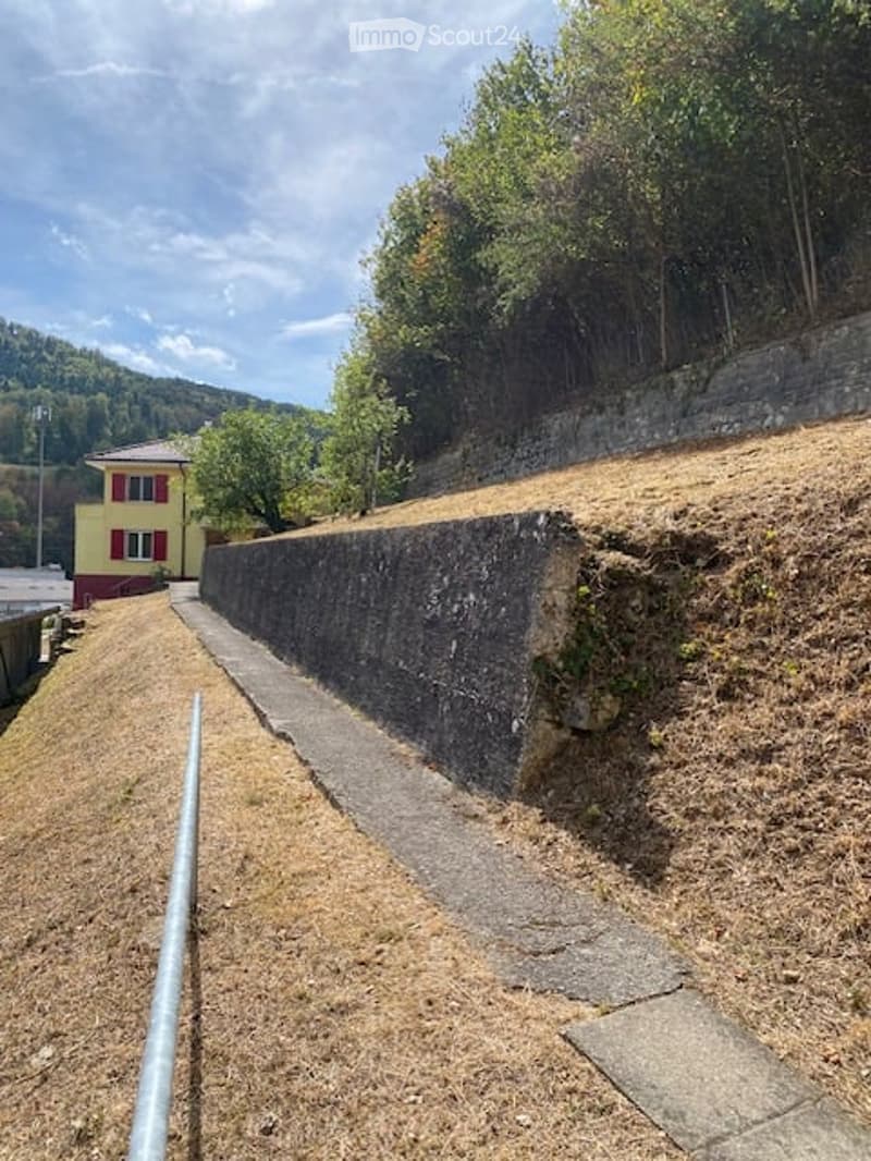 A raised ground floor pathway bordered by a stone retaining wall, with grass and trees on the hillside above.