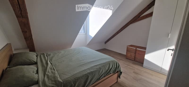 Cozy attic bedroom with wooden bed, green bedding, and two wicker storage boxes. Slanted wooden beams and skylight provide natural light.