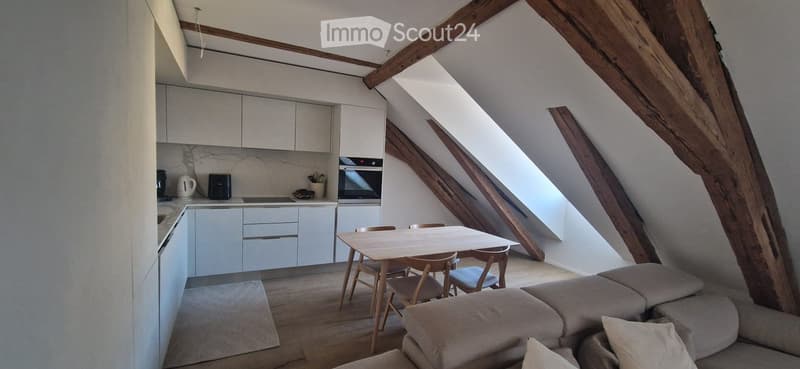 Modern attic apartment with exposed wooden beams, a sofa, a dining table with chairs, and a fitted kitchen area with cabinets and appliances.