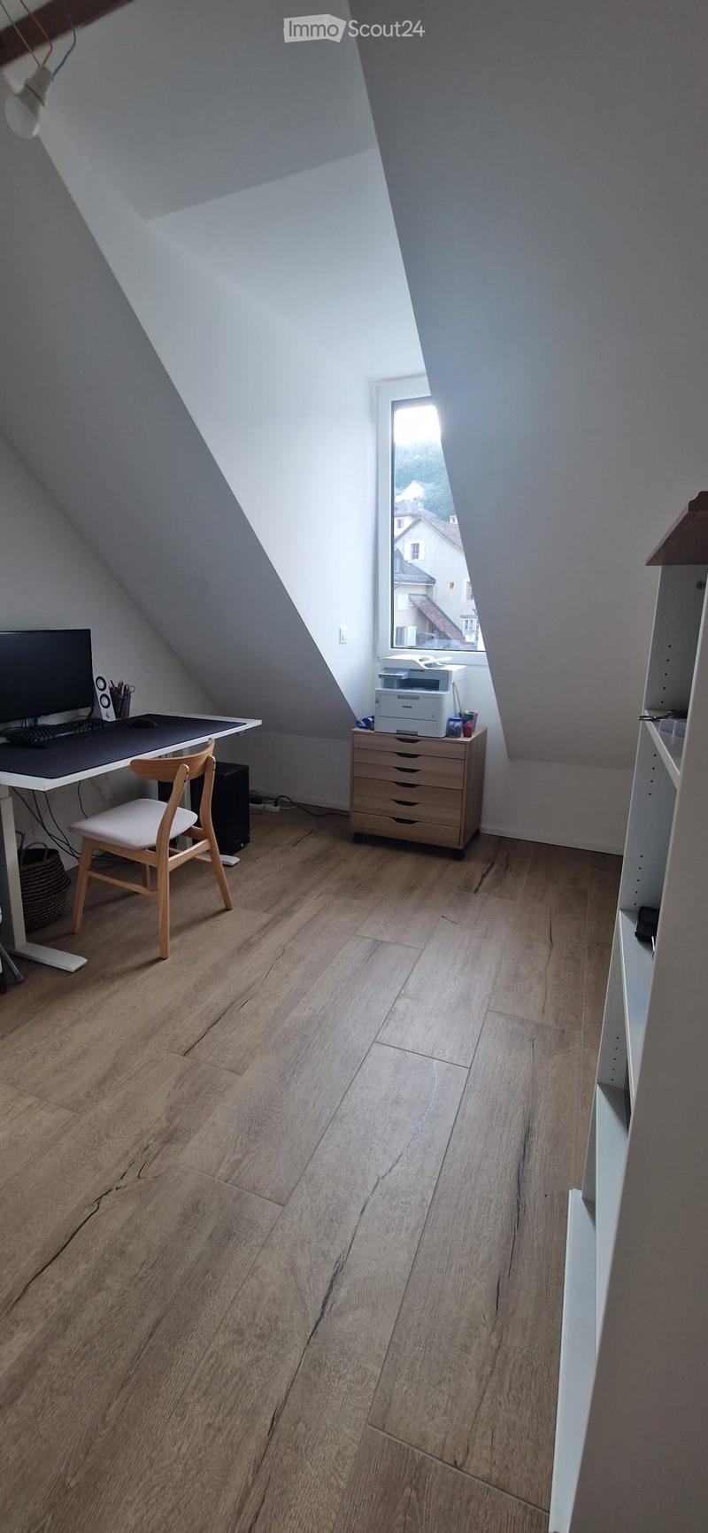 Bright attic room with wooden flooring, slanted ceilings, and large windows. Contains a desk with a computer, chair, and printer on a small cabinet.