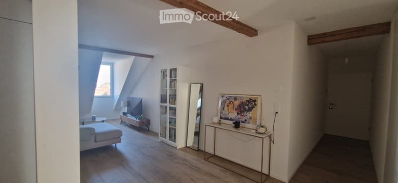 Bright living room with wooden floors, white walls, and exposed wooden beams. Features a white sofa, glass coffee table, bookshelf, mirror, and a small table with decorative items.