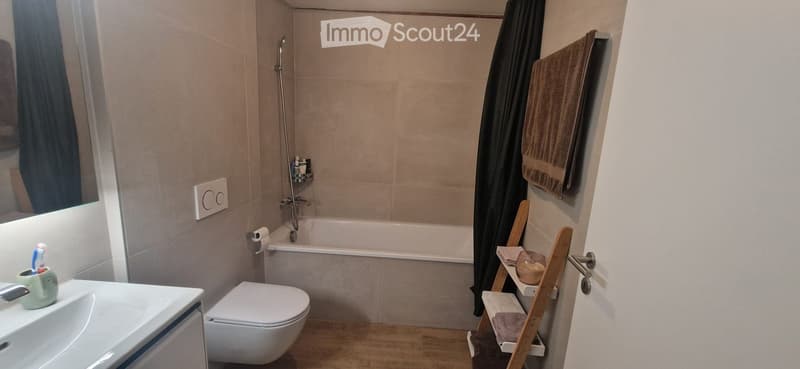 Modern bathroom with a white sink, toilet, bathtub, and a wooden shelf holding towels. A mirror is mounted on the wall, and a toothbrush holder sits on the sink.