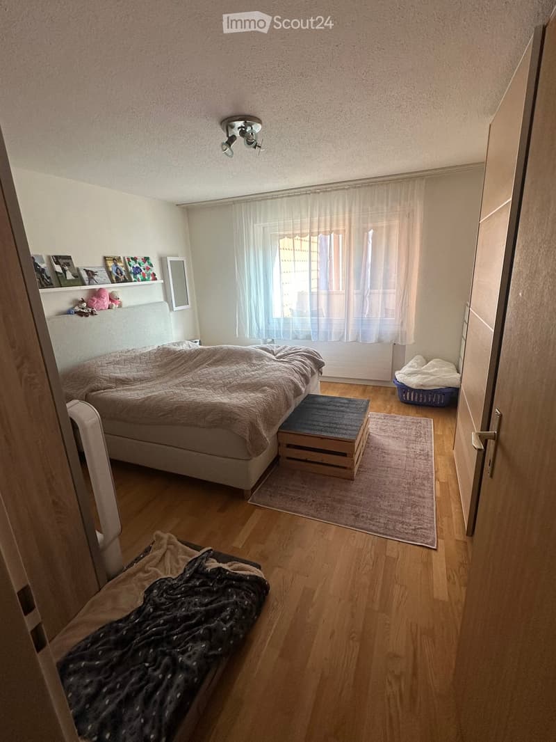 Cozy bedroom with wooden flooring, a large bed with a headboard, a nightstand, a small stool, a mirror, a shelf with decorative items, and a window with curtains.