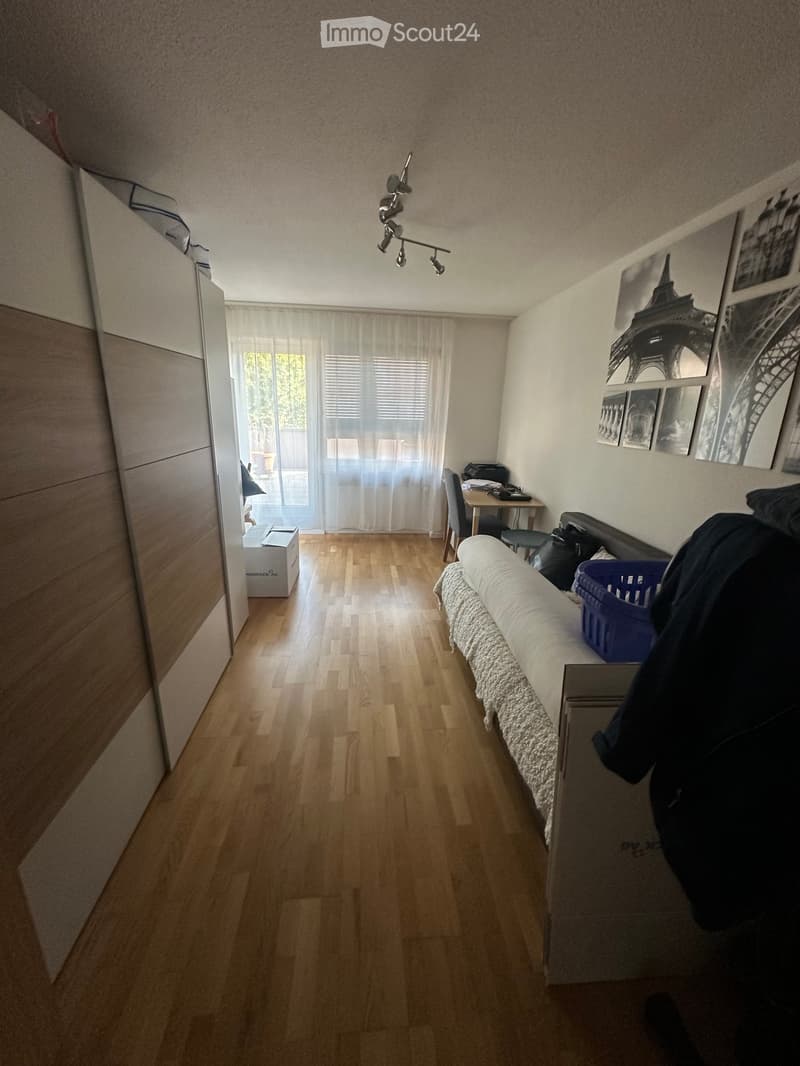 A furnished room with a sofa, a small table, a chair, a wardrobe, and a window with curtains. The room has wooden flooring and several picture frames on the wall.