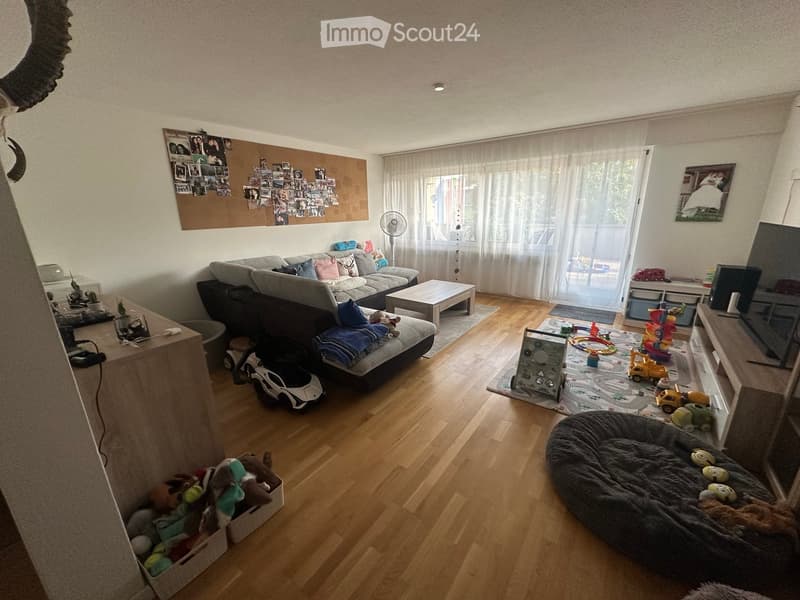 Living room with wooden flooring, large window with white curtains, grey sectional sofa with pillows, coffee table, dog bed, toys, and a fan. There is a bulletin board with photos on the wall and a TV on a stand.