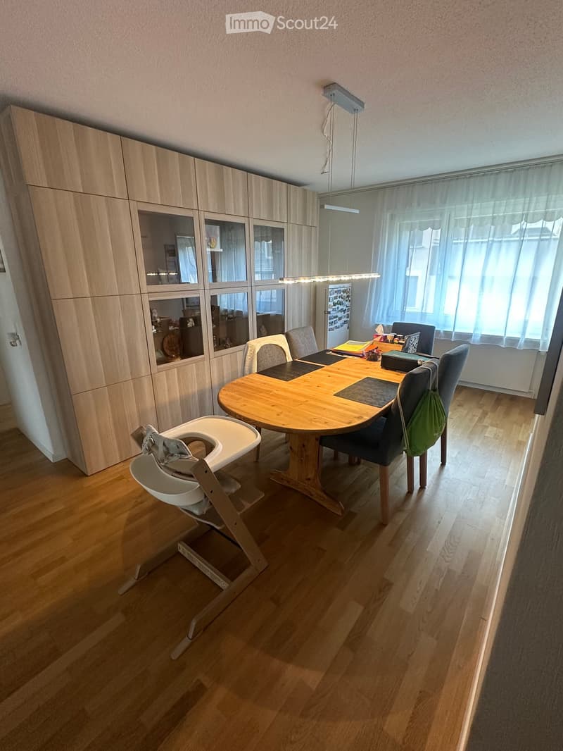 Dining area with a wooden table, chairs, a baby chair, and a large wooden cupboard with glass panels.