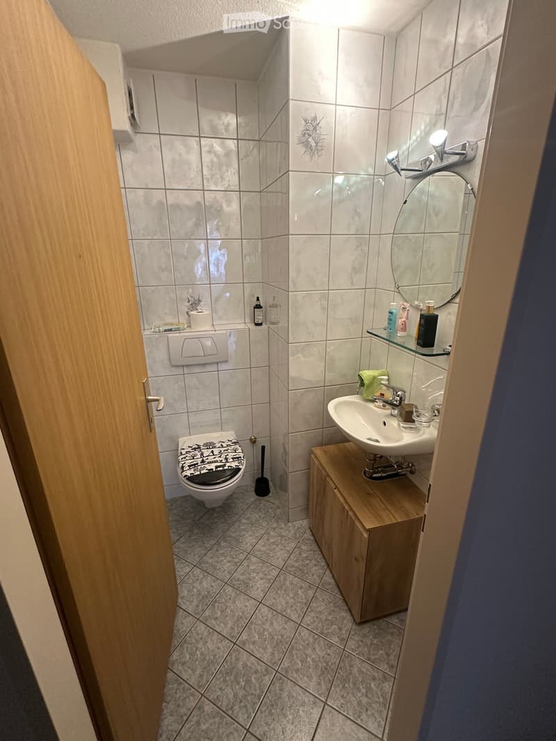 Modern bathroom with white tiled walls, a toilet with a patterned seat cover, a sink with a wooden cabinet, a mirror, and a glass shelf holding toiletries.