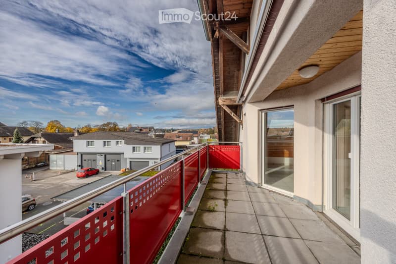 Balcony with a red metal fence, tiled floor, and a view of a residential area with houses, a parking lot, and a clear sky.