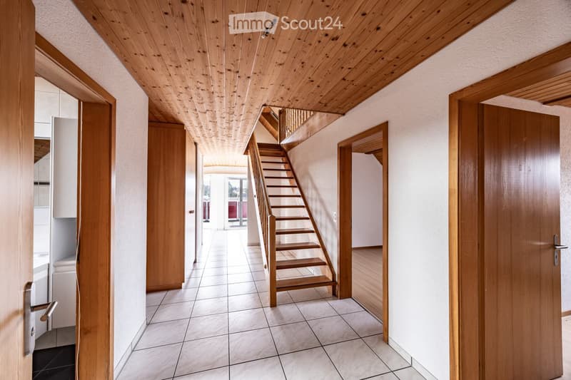 Bright hallway with wooden ceiling, tiled floor, and wooden staircase leading upwards. Doors on both sides lead to other rooms.