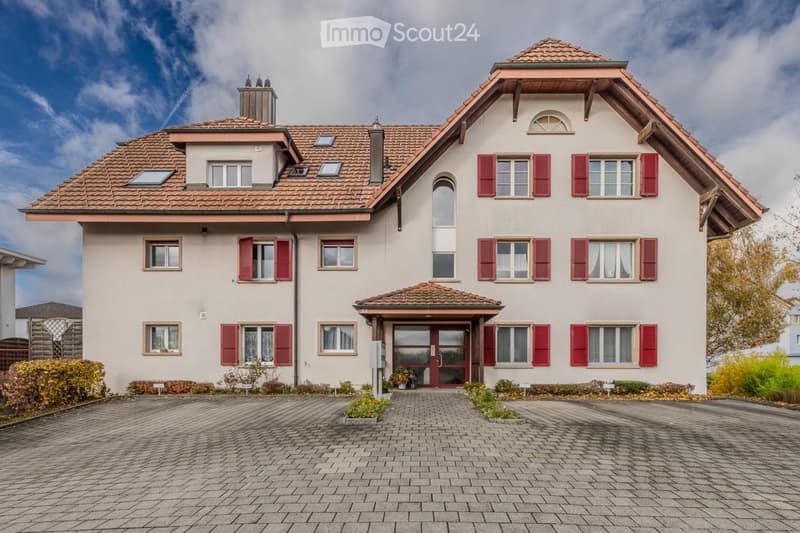 Two-story house with red shutters, beige walls, tiled roof, and a brick driveway.