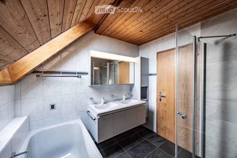Modern bathroom with a wooden slanted ceiling, white tiled walls, a bathtub, a double sink with a mirrored cabinet, and a glass-enclosed shower area.