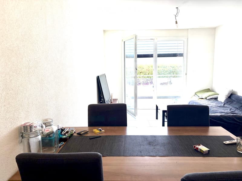A modern, well-lit room with a large window and sliding glass doors leading to a balcony. The room features a wooden dining table with chairs, a sofa, and a television. There are various items on the table, including a jar, a cup, and some small objects.